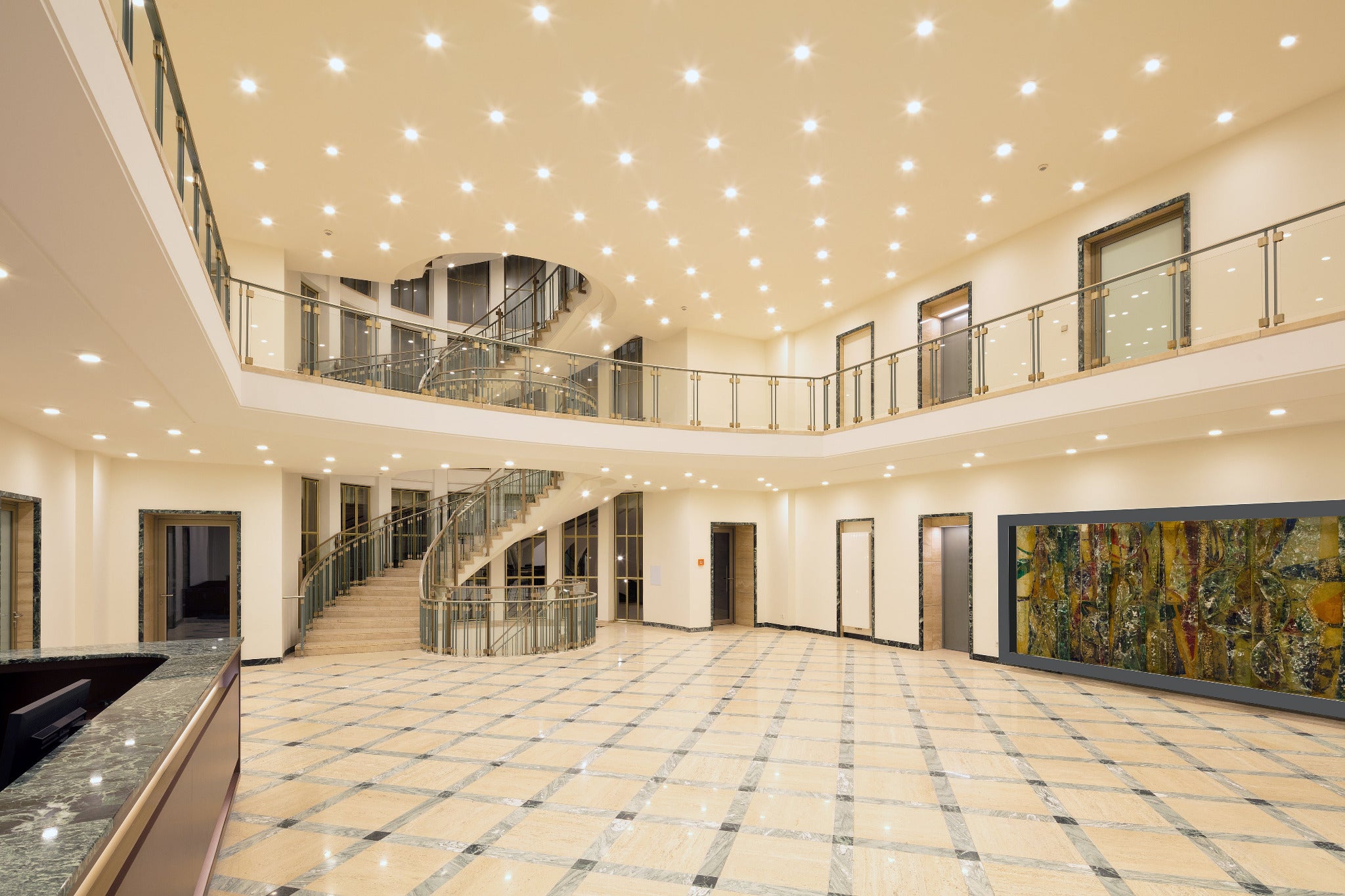 Innenbereich mit breiter Treppe, Glasgeländern, Marmorboden und zahlreichen Deckenleuchten in hellem Foyer